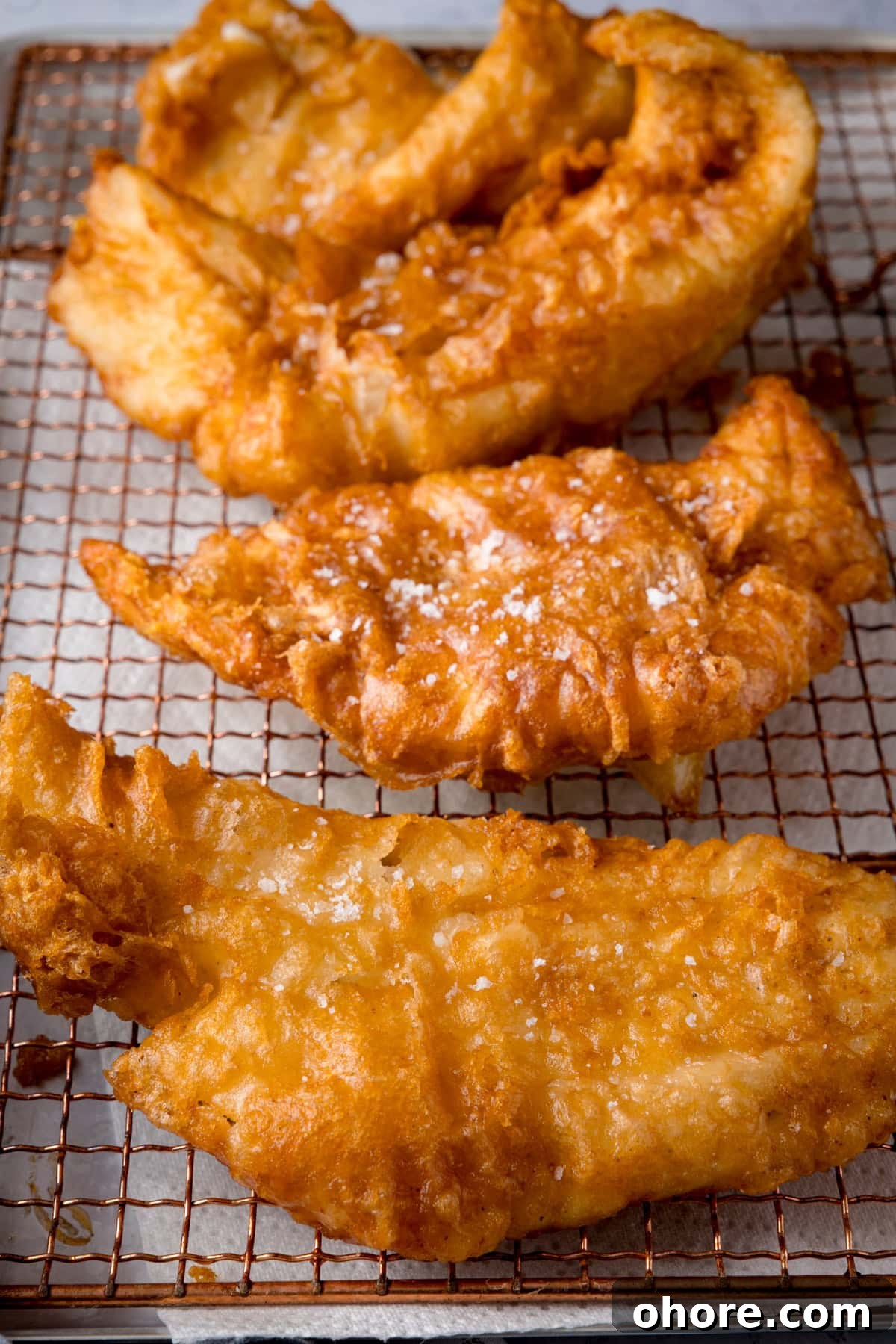 A tall shot of three fish fillets, battered in the beer batter. There are three golden battered fish fillets laid side by side on a copper wire rack. They are topped with flakes of salt. The wire rack is on a white marble background.
