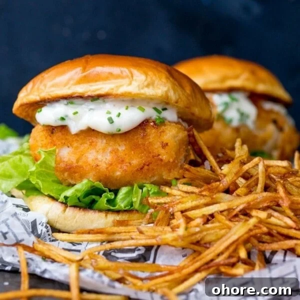 Breaded haddock burger on brioche bun with lettuce and chive-speckled mayonnaise. Shoestring fries to the right of the burger. A second burger in background.