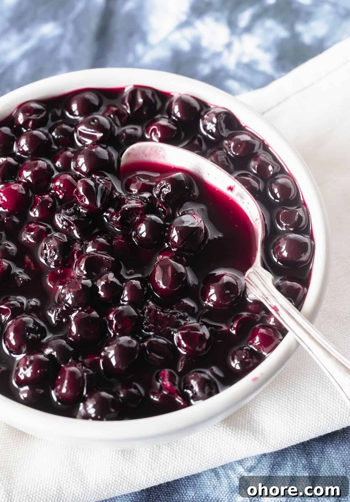 Blueberry Compote Recipe: Simple Homemade Blueberry Sauce 2 Partial view of white bowl with silver spoon and blueberry compote on white blue cloth.
