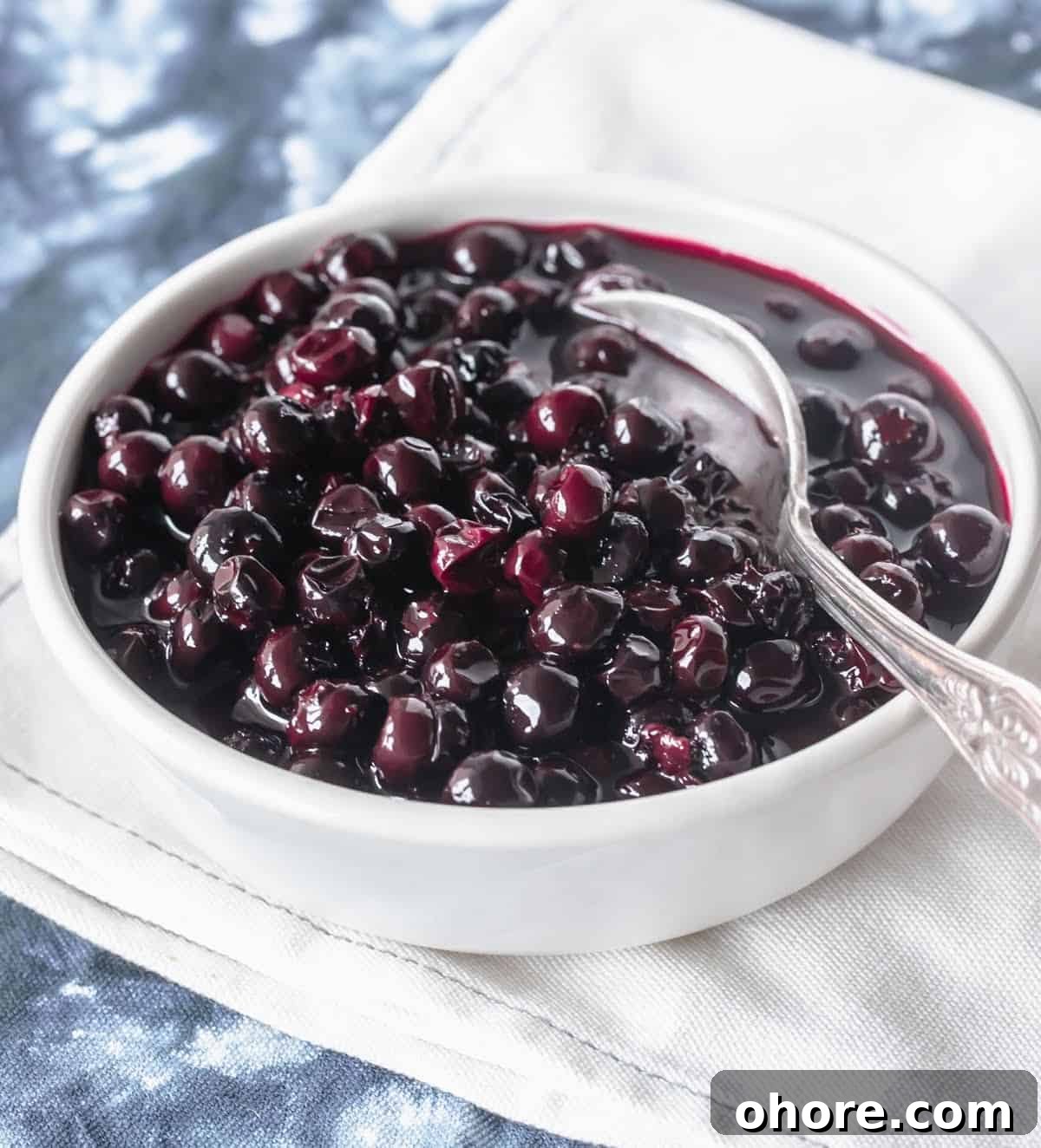Blueberry Compote Recipe: Simple Homemade Blueberry Sauce 3 Whole white bowl with blueberry compote and silver spoon on blue white cloth.