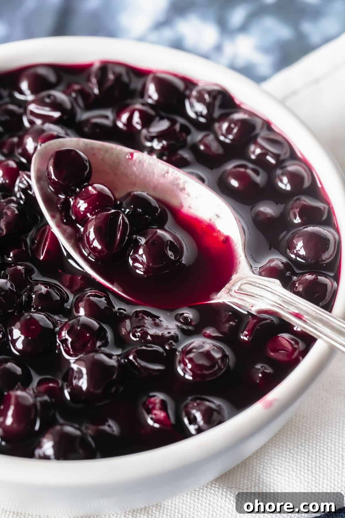 Blueberry Compote Recipe: Simple Homemade Blueberry Sauce 7 Silver spoon with blueberry compote over white bowl with compote. Blue white background.