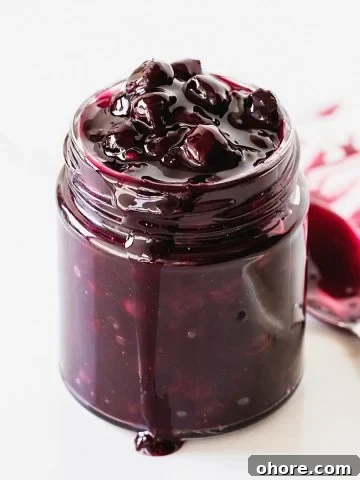 Glass jar with blueberry sauce, a spoon, white background.
