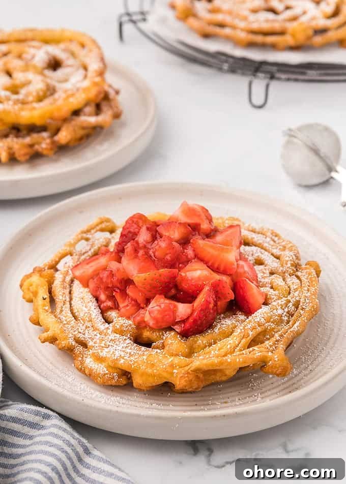 Crispy Homemade Funnel Cake Recipe for Fair-Style Treats 8 Homemade Funnel Cakes topped with powdered sugar and strawberries on a white plate on a marble surface with a blue striped linen next to it