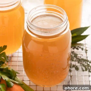 Golden chicken bone broth in glass jars with carrots, celery, and parsley, cooling on the counter.