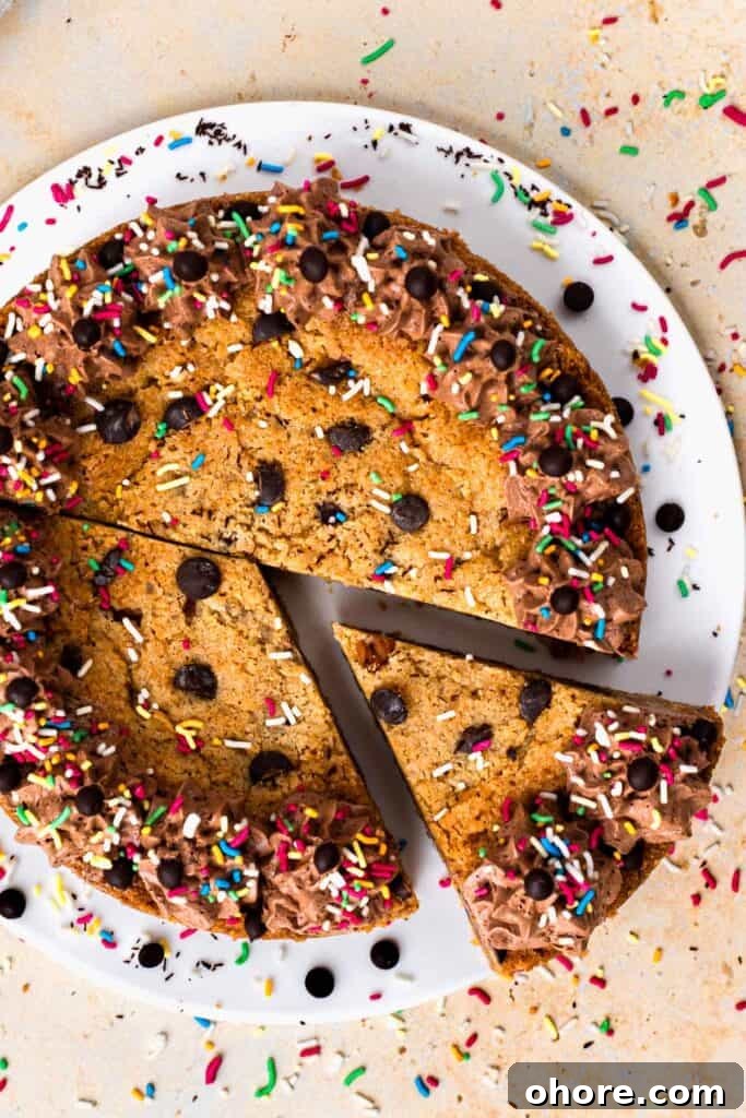 Gluten-Free Cookie Cake Recipe: Soft, Chewy Sugar Cookie Cake 2 Top view of a gluten free cookie cake with a slice taken out, topped with chocolate buttercream on the outside, sprinkles and chocolate chips, sitting on a white plate, and surrounded by more sprinkles.