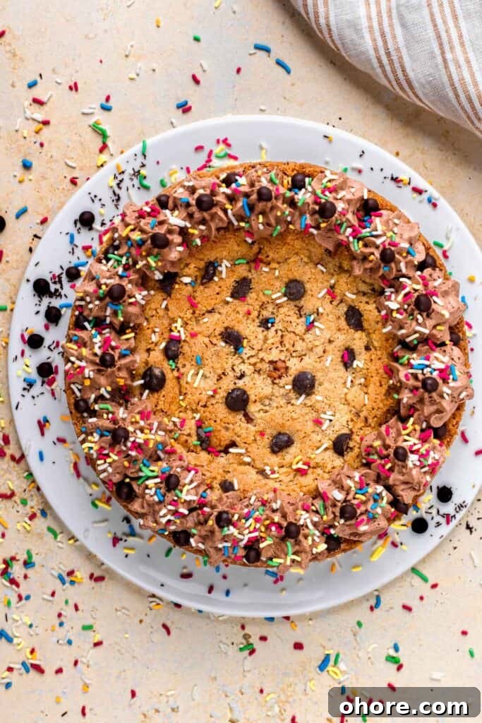 Gluten-Free Cookie Cake Recipe: Soft, Chewy Sugar Cookie Cake 7 Top view of a gluten free cookie cake topped with chocolate buttercream on the outside, sprinkles and chocolate chips, sitting on a white plate, and surrounded by more sprinkles.