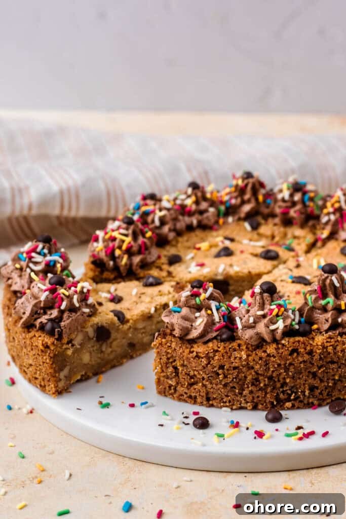 Gluten-Free Cookie Cake Recipe: Soft, Chewy Sugar Cookie Cake 8 Side view of a gluten free cookie cake with a piece cut out, topped with chocolate buttercream on the outside, sprinkles and chocolate chips, sitting on a white plate, and surrounded by more sprinkles.