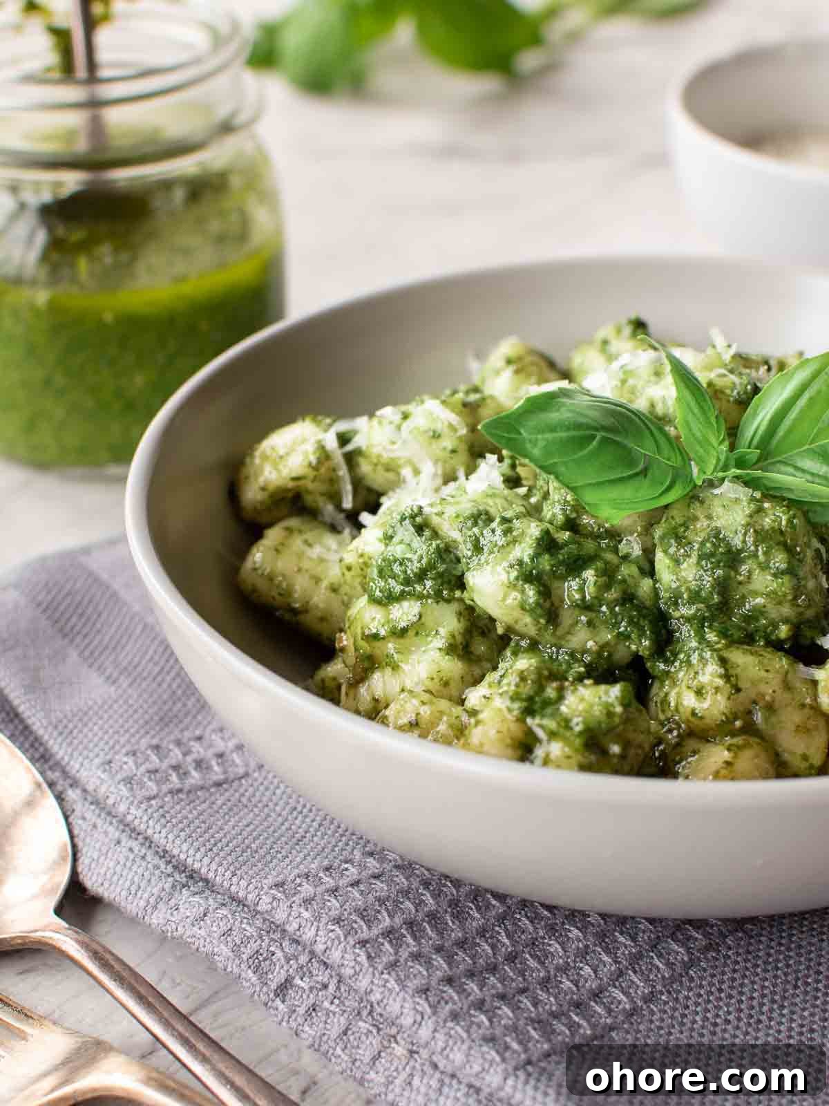 Creamy Pesto Gnocchi with Parmesan and Basil 2 A bowl this pesto gnocchi recipe on a grey cloth with jar of pesto in the background.