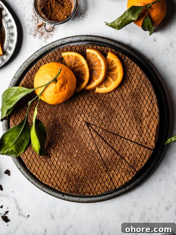 A chocolate orange cheesecake decorated with orange slices and a whole orange with a few slices removed, on a white marble surface.