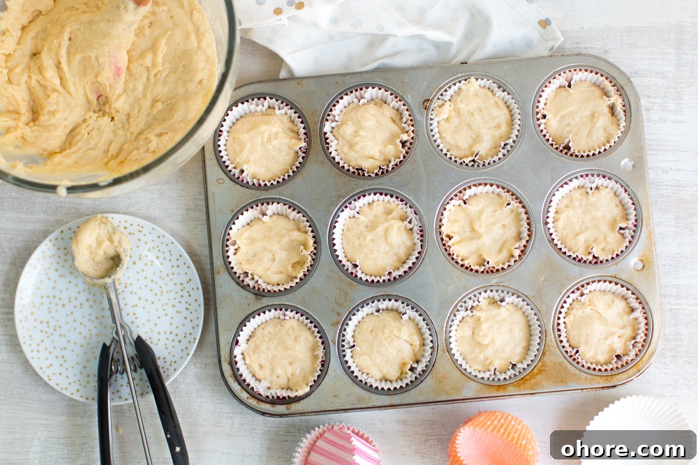 a cookie scoop sits beside a bowl of cupcake batter and 12, filled cupcakes liners in a tray ready for the oven