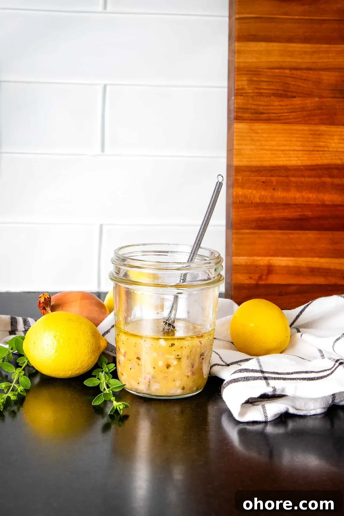 Yellowish salad dressing in a jar with a whisk and some lemons and fresh herbs surrounding on the counter.