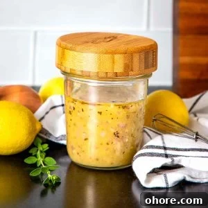 Glass jar with wooden lid filled with salad dressing, surrounded by fresh lemons, a shallot and some fresh herbs.