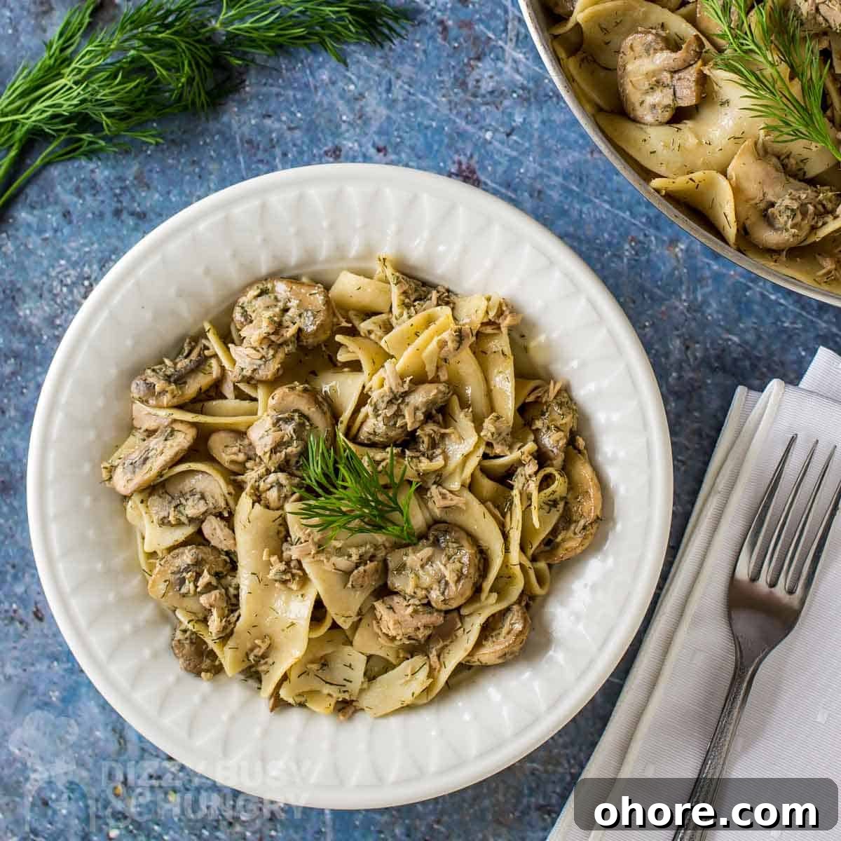 Quick Weeknight Dinners When You're Short on Time and Supplies 4 Overhead shot of lemon dill tuna and noodles garnished with herbs in a white bowl on a blue placemat with herbs and silverware on the side.