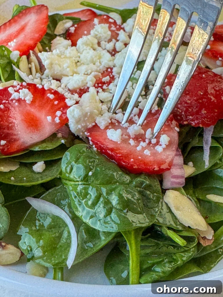 Close up fork in a strawberry slice with crumbled feta over a bed of baby spinach with sliced almond and red onions.