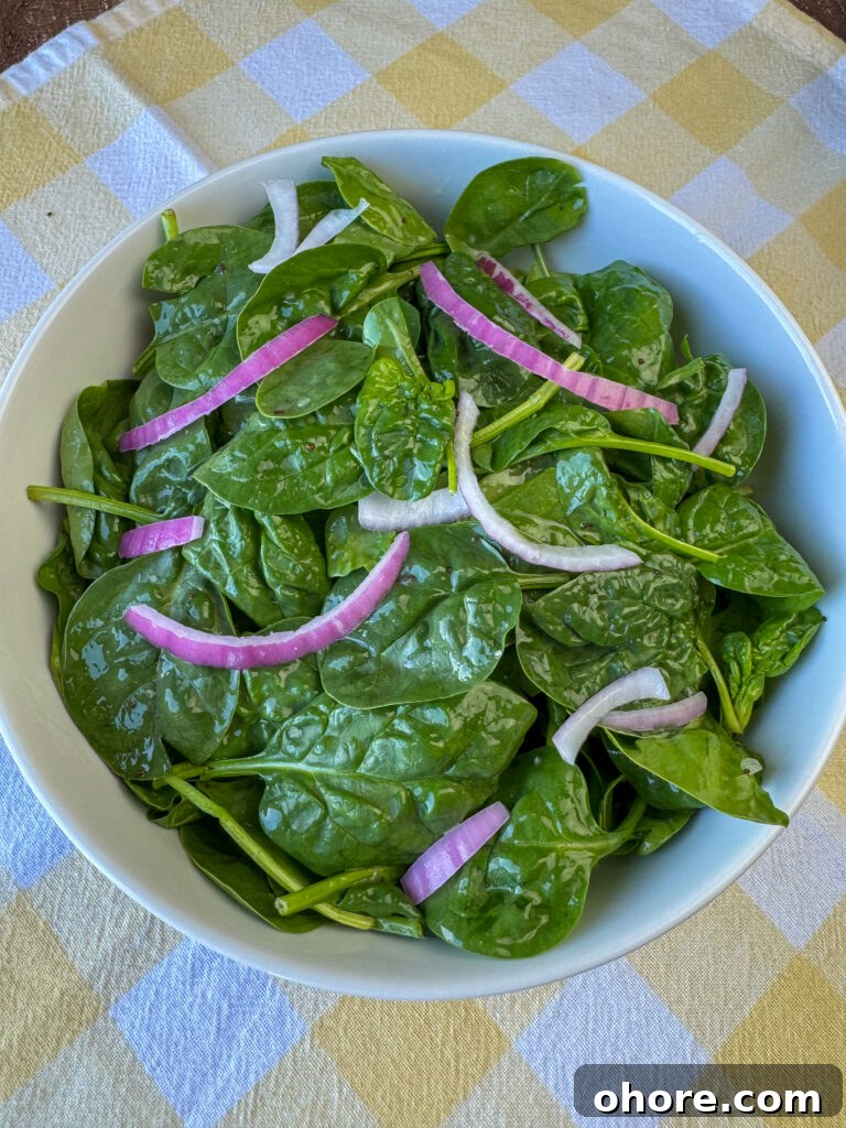 Sliced red onions on top of baby spinach with dressing in a white bowl.
