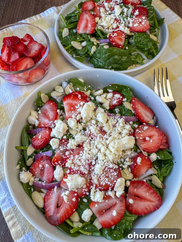 Overhead view of a generous salad with feta cheese sprinkled over strawberries, a few red onion slices and lots of baby spinach in a bowl with another plate of salad, more sliced berries and a fork on a yellow and white napkin surrounding the bigger white bowl full.