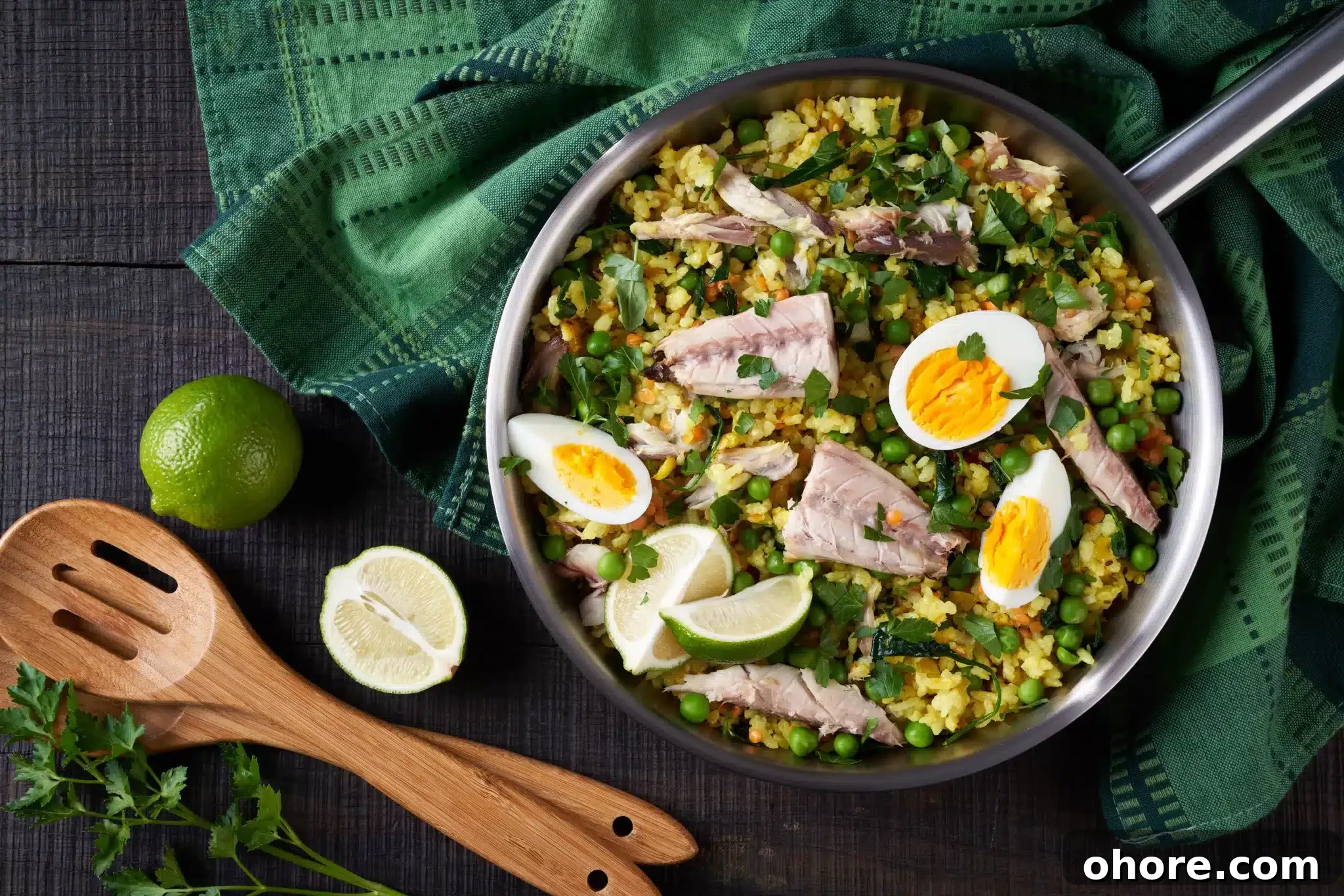 Top view of a dark table displaying a pan with kedgeree, lime, parsley, and a couple of wooden spoons.