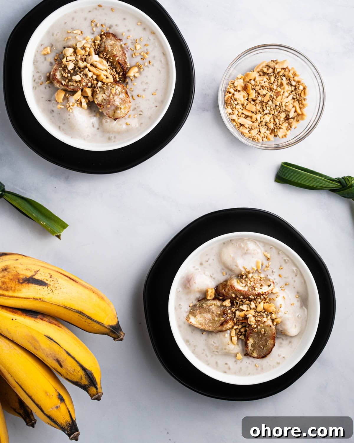 two bowls of che chuoi surrounded by bananas, pandan leaves tied in knots, and a bowl of crushed peanuts and sesame seeds.