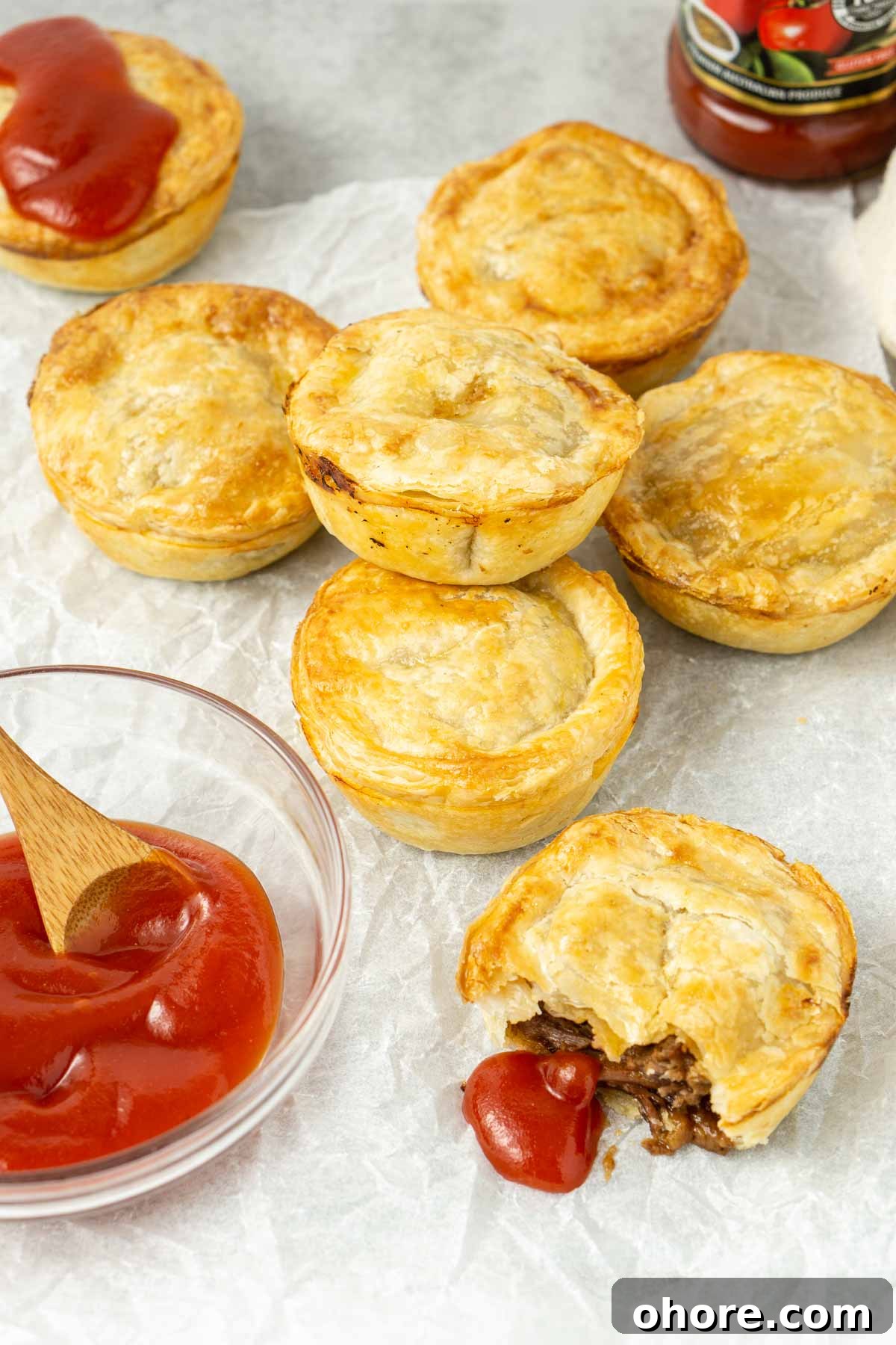 Mini beef pies laid out on baking paper with tomato sauce.
