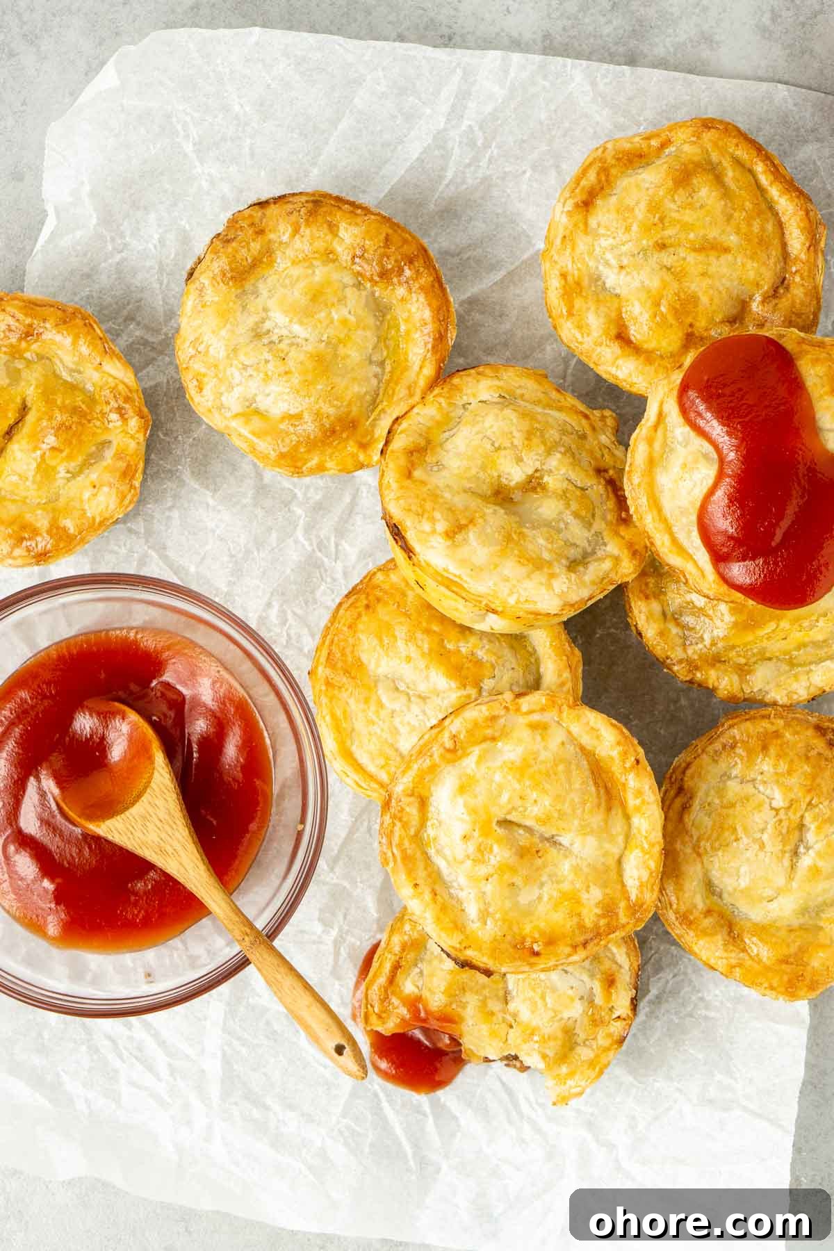 Mini beef pies laid out on baking paper with tomato sauce.