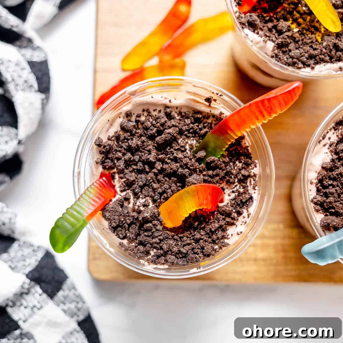 Kid-Friendly Halloween Snack Ideas for Kids 11 A cup filled with chocolate pudding, crumbled cookies, and gummy worms on top, placed on a wooden board next to a black and white checkered cloth.