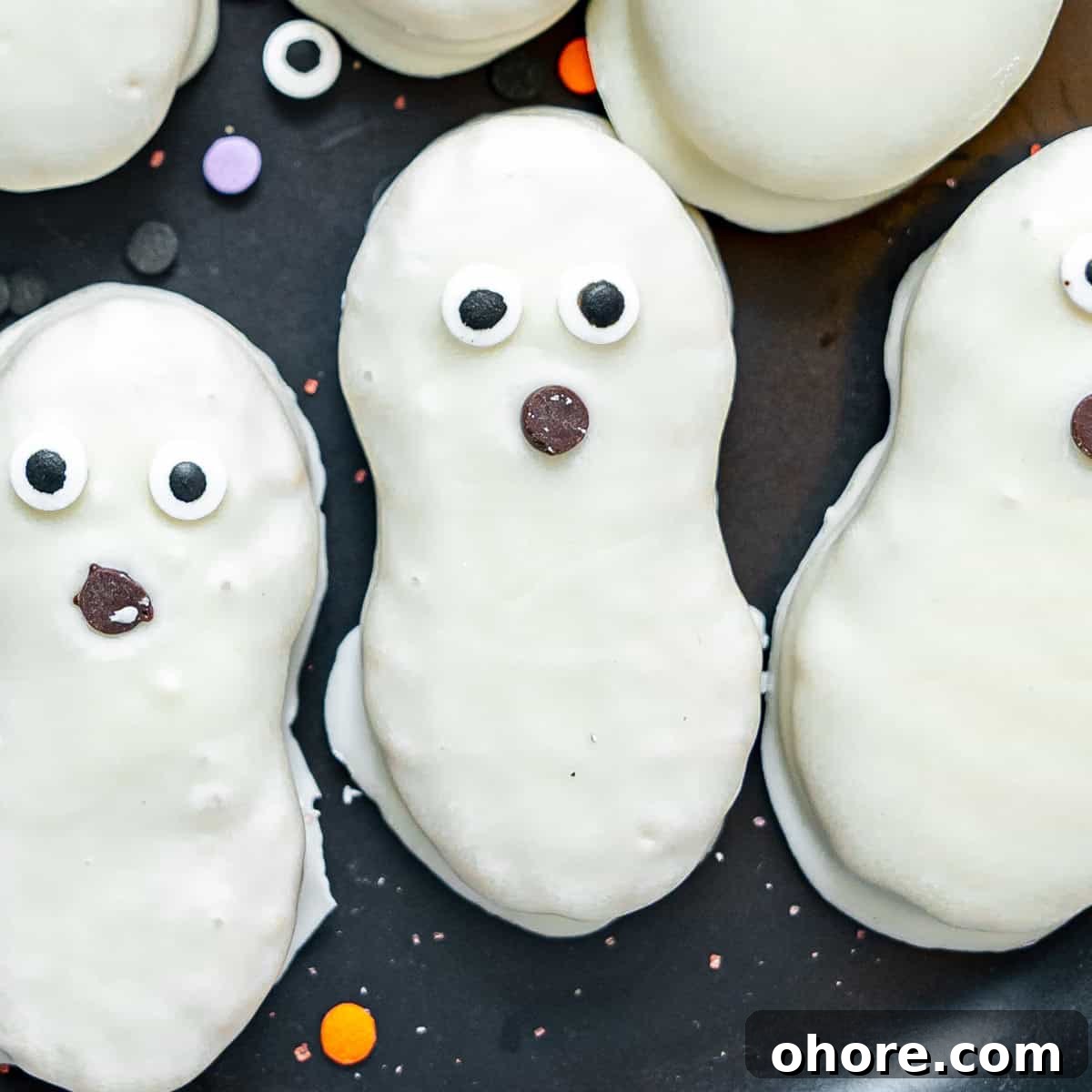 Kid-Friendly Halloween Snack Ideas for Kids 10 Three ghost-shaped cookies with white icing, candy eyes, and chocolate chip mouths are arranged on a black surface with colorful round sprinkles.