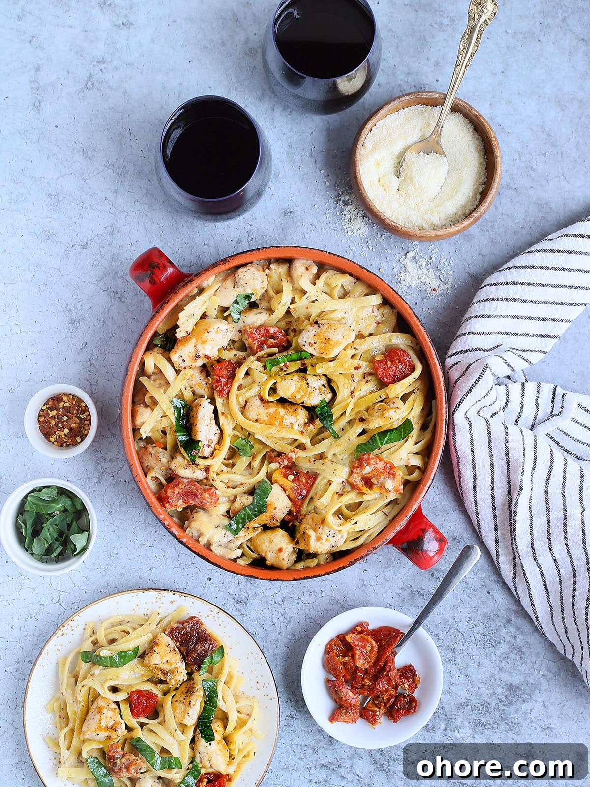 Creamy Marry Me Chicken Pasta Recipe for Weeknight Dinner 2 Serving bowl of Marry Me Chicken Pasta with two glasses of red wine, small dishes of grated Parmesan and sundried tomatoes, and an individual plate of pasta on the side.