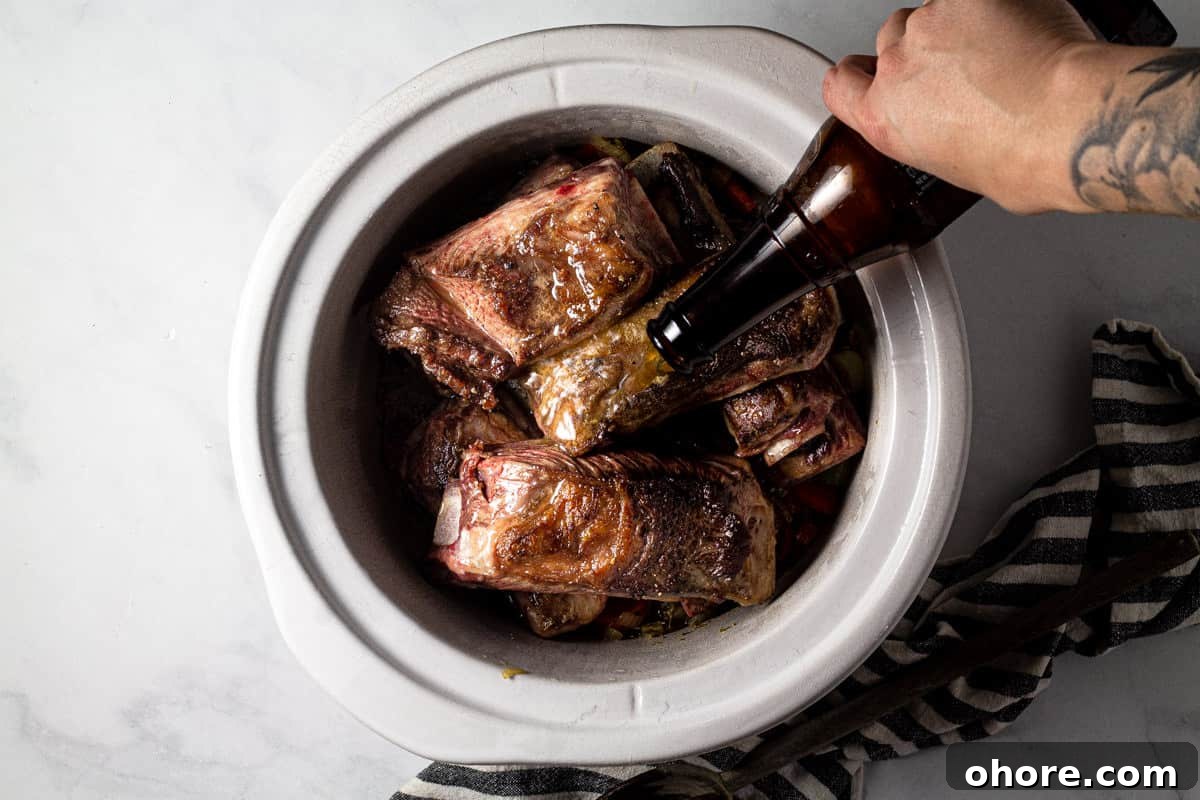 Large crock pot insert filled with seared short ribs with beer being poured over them