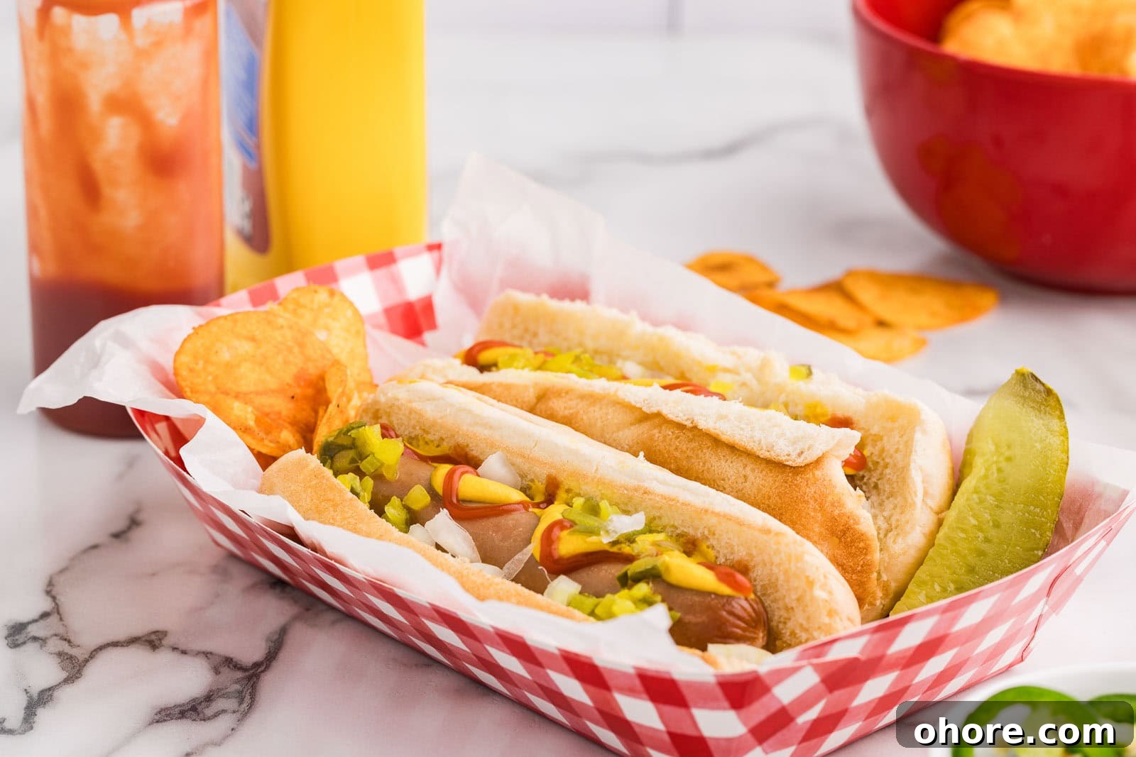 Slow Cooker Hot Dogs: Juicy, Fuss-Free Recipe and Tips 6 red and white paper tray with 2 dressed hot dogs, potato chips and dill pickle with bottle of ketchup and mustard, red bowl of potato chips in background
