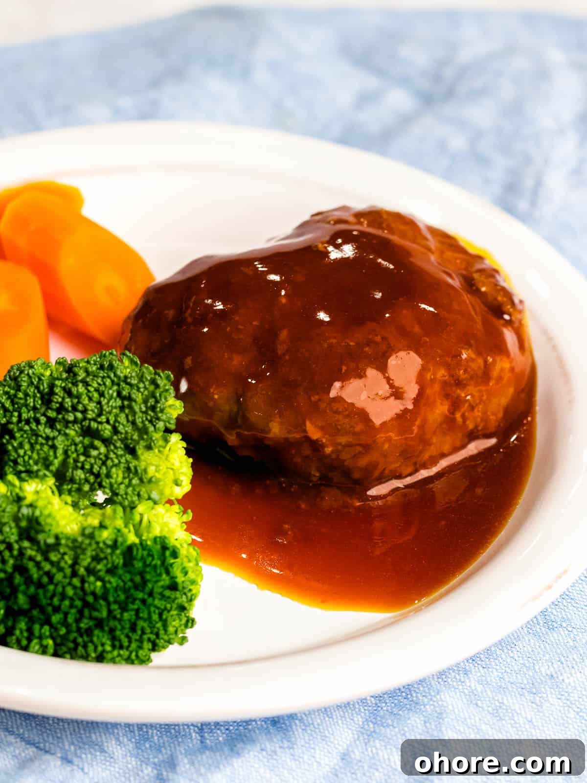 Japanese Hambagu (Hamburger Steak) Recipe — Authentic Pan-Fried Steak 3 Juicy Japanese hamburger steak (hambāgu ハンバーグ) covered in sauce on a white plate next to carrots and broccoli.