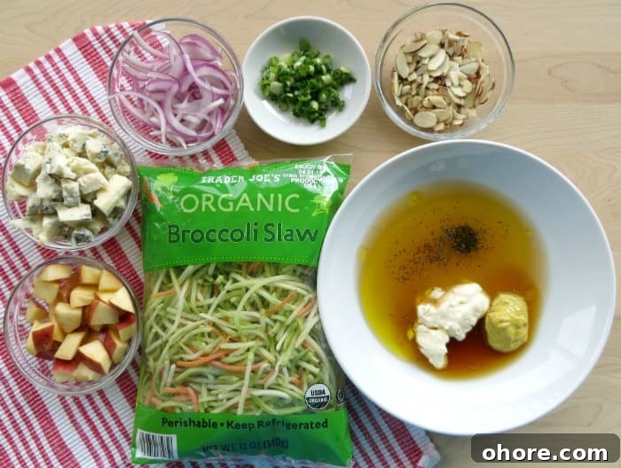 Quick Broccoli Slaw Recipe You Can Make Tonight 3 Broccoli Slaw ingredients on a wood cutting board, apples, blue cheese, red onions, slivered almonds, and more